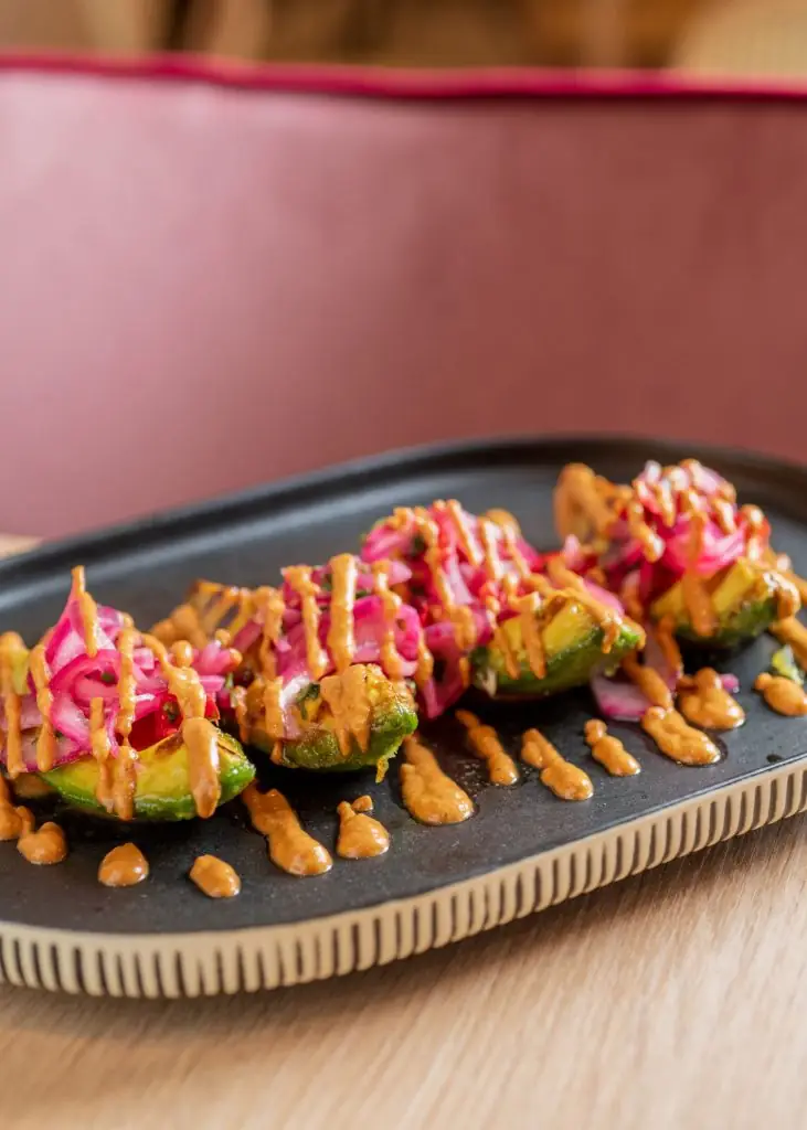 Black oval plate of grilled avocado halves topped with pickled red onion and drizzled with a brown romesco-style sauce, on a wooden table