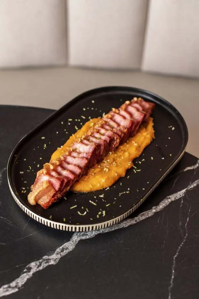 Sliced roasted pork belly arranged in a row over an orange squash puree with lime zest, on a black oval plate on marble