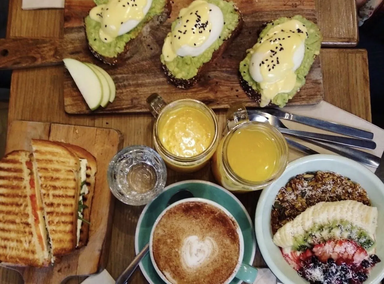A wooden table with avocado toasts, a toasted sandwich, a latte, orange juices and a fruit bowl