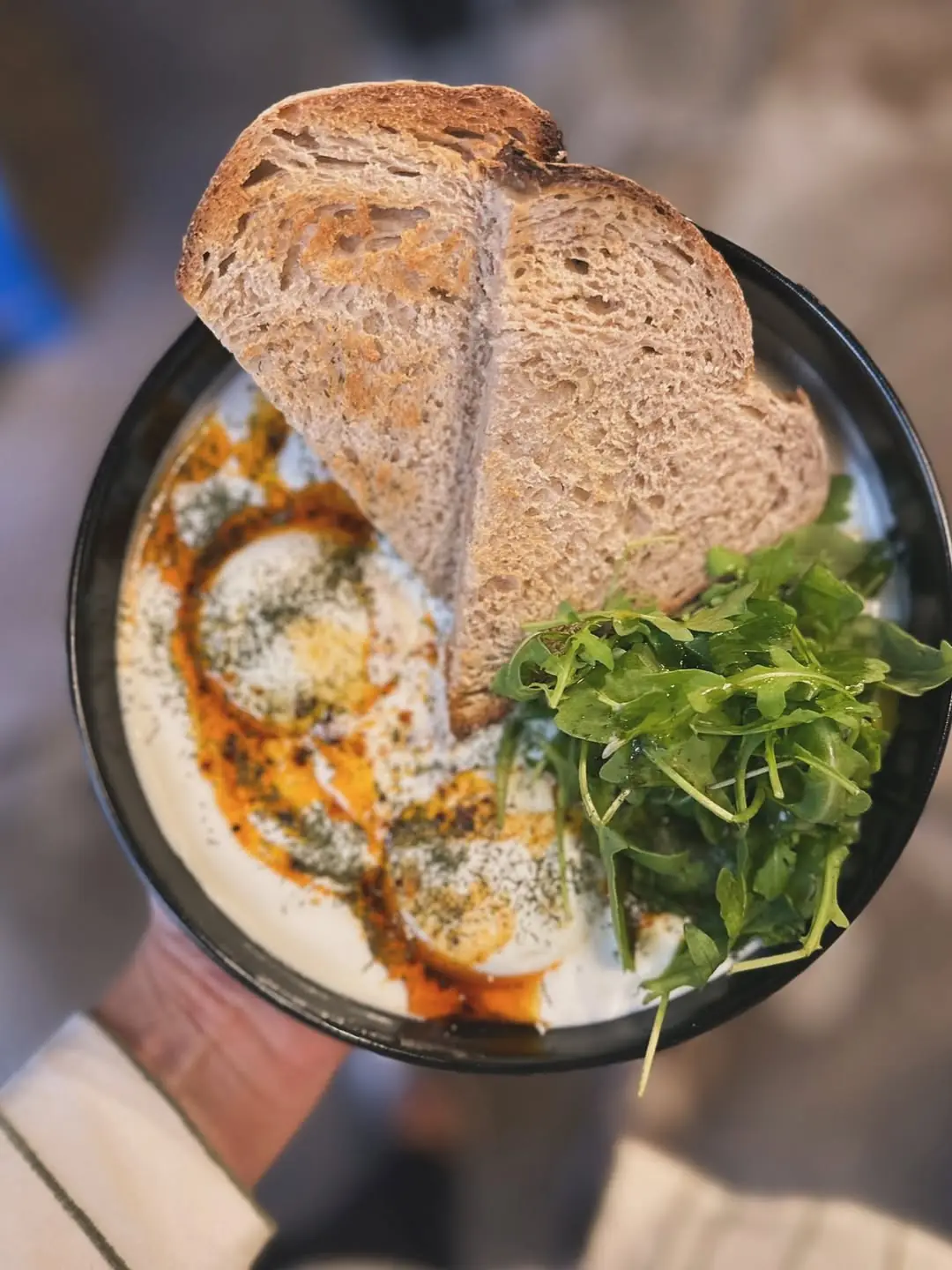 An overhead shot of baked eggs with toast and arugula salad, held in hand