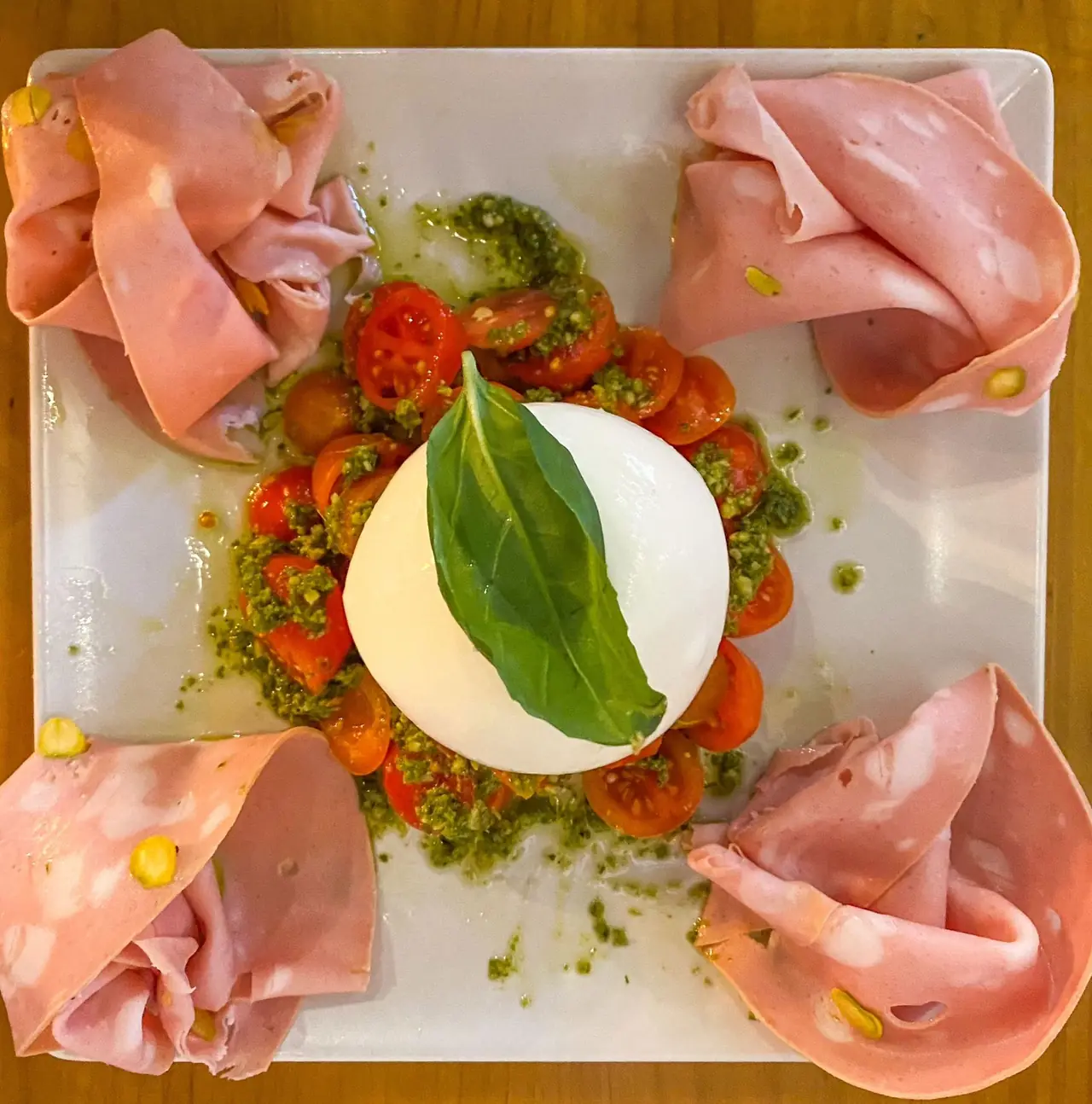 Whole burrata topped with a basil leaf, surrounded by cherry tomatoes in pesto and folded slices of mortadella on a white plate at Pummarola Pizzeria Napoletana