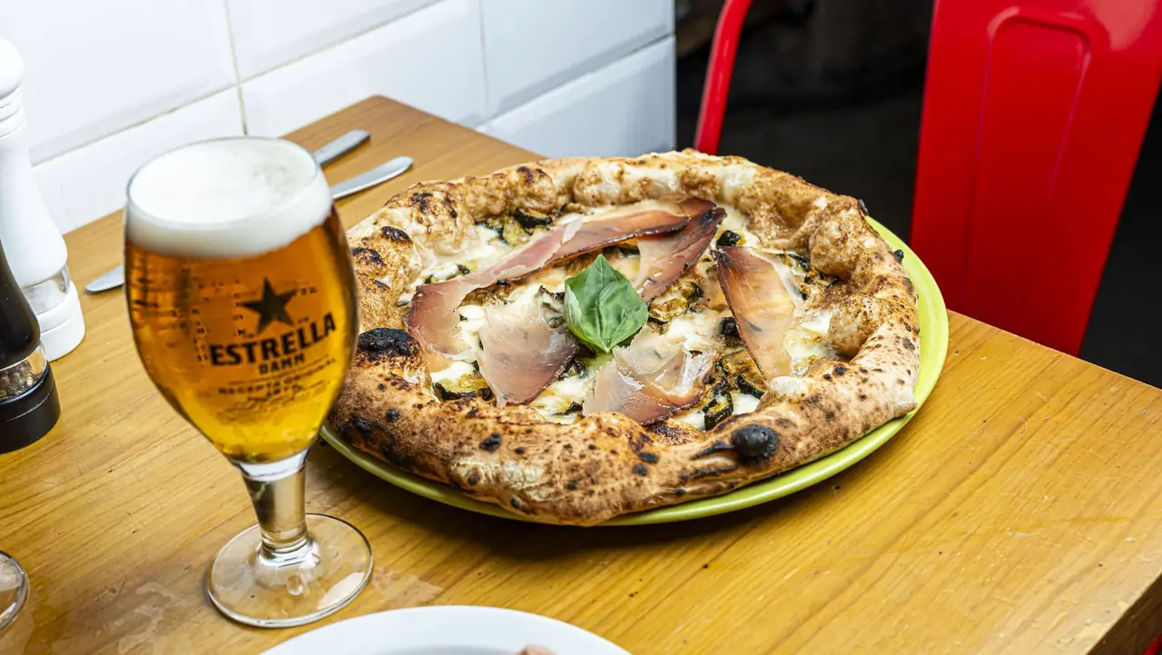 Wood-fired pizza with melted cheese, zucchini, thin slices of cured ham, and a basil leaf, served with a glass of beer at Pummarola Pizzeria Napoletana