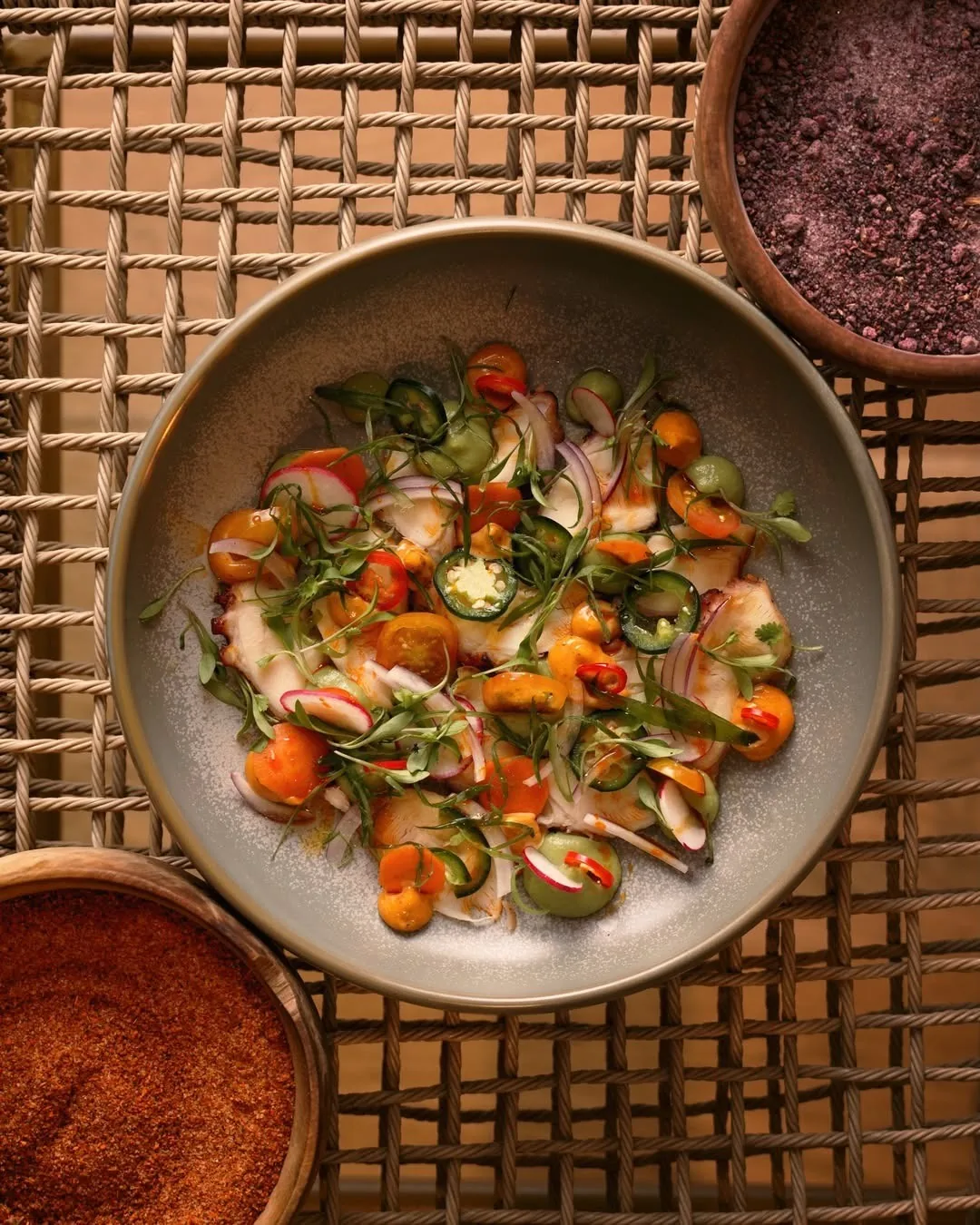 A bowl of sliced seafood with chilli slices, herbs and cherry tomatoes on a woven mat