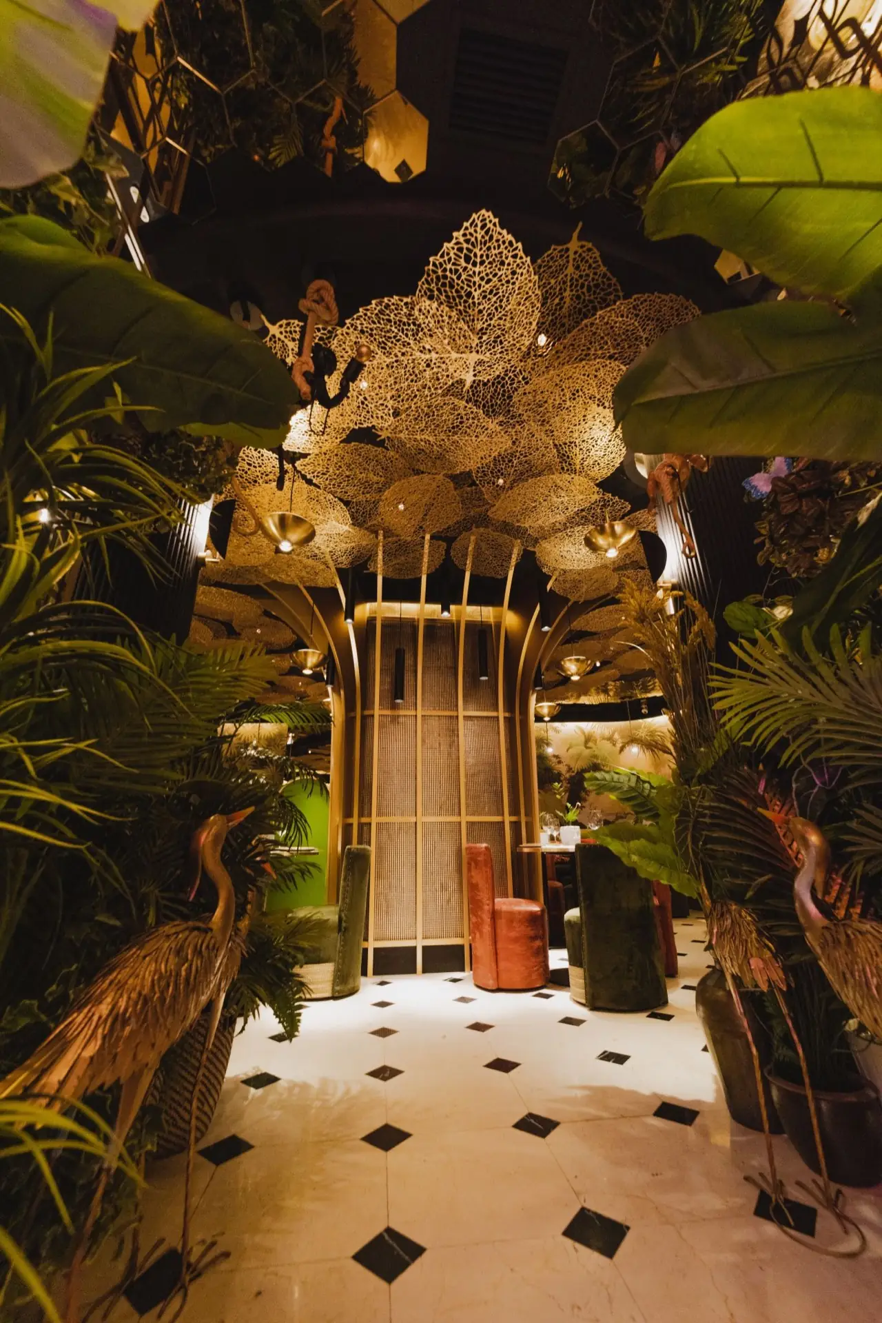 Dramatic restaurant entrance with a gold filigree leaf-canopy ceiling, lush tropical plants, gilded crane sculptures and green and rust velvet stools on a black-and-white diamond tile floor