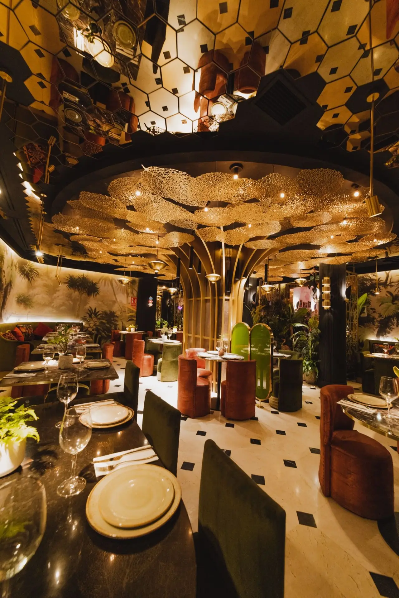 Jungle-themed dining room with a golden filigree cloud ceiling, mirrored hexagon panels, rust and green velvet armchairs and set tables on diamond-patterned marble flooring