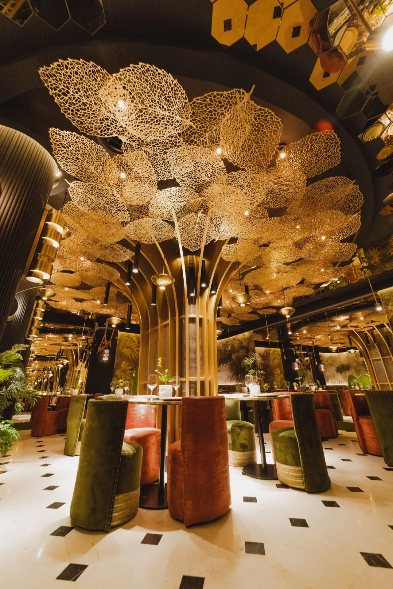 Central golden tree-shaped column blossoming into a gilded lace-leaf ceiling canopy over green and terracotta velvet tub chairs and marble bistro tables