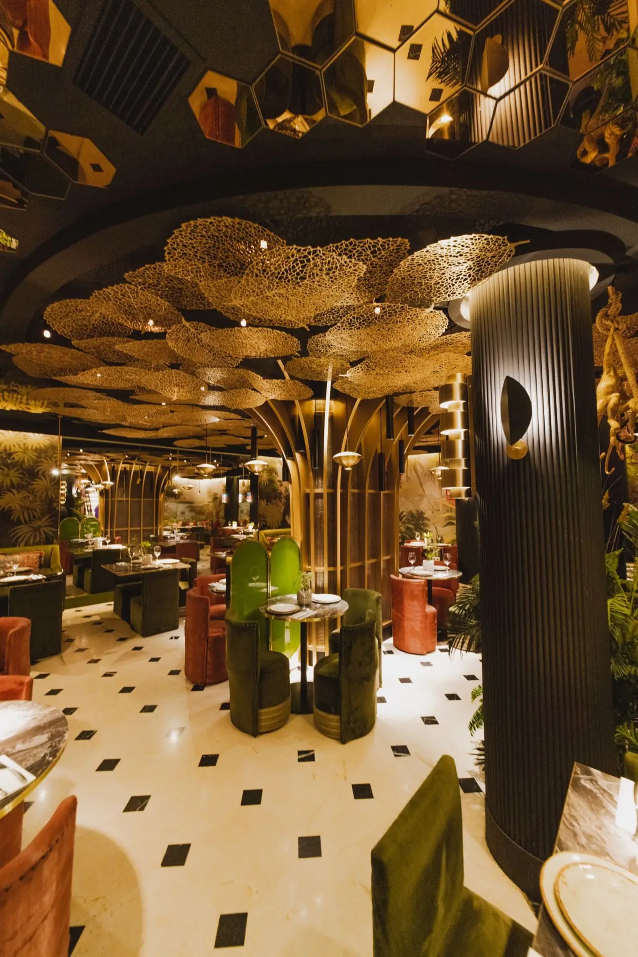 Ornate dining room with gilded leaf ceiling sculpture, mirrored hexagon ceiling tiles, a fluted black column and green and rust velvet chairs at set marble tables