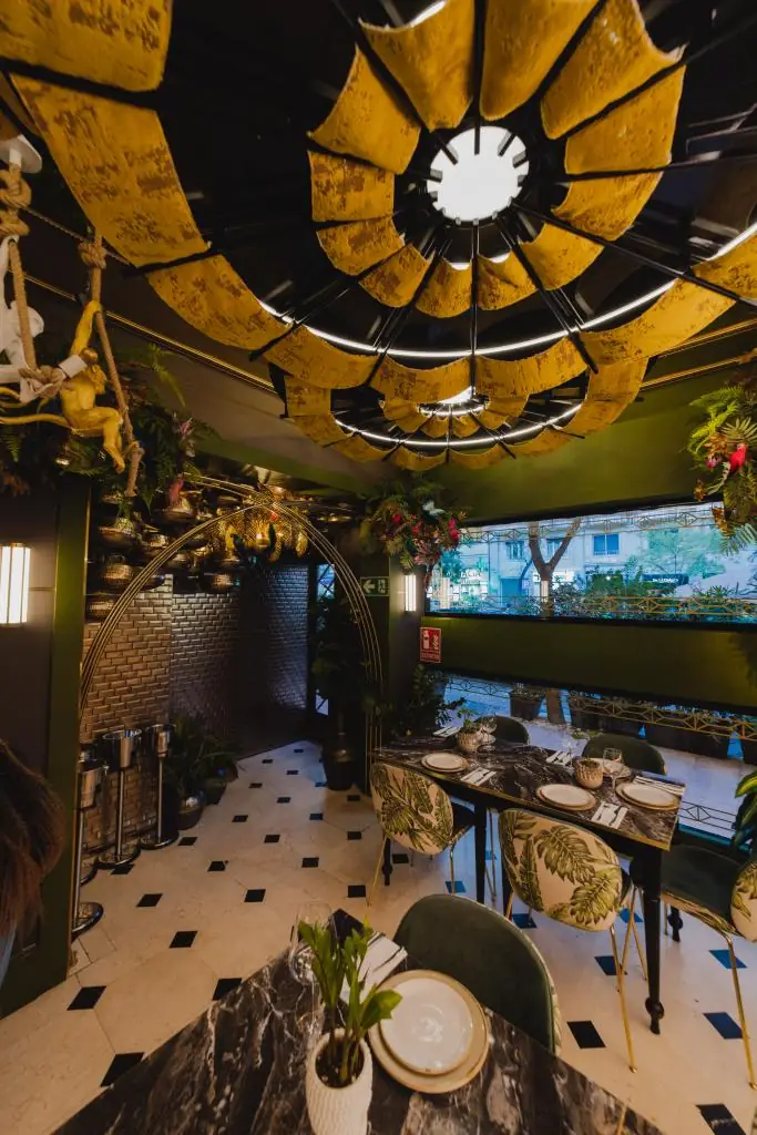 Intimate dining nook with a scalloped gold ceiling installation, botanical-print upholstered chairs, marble tables and a window looking onto a tree-lined street
