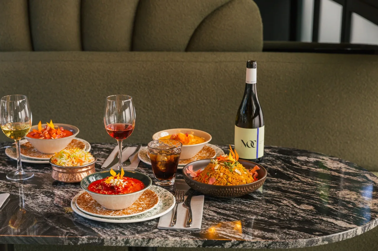 A marble table set with bowls of orange and red curries, saffron rice and a copper dish of white rice beside wine glasses and a bottle
