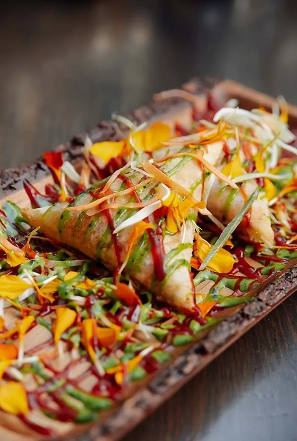 Two crisp triangular pastries on a bark-edged board scattered with edible petals and drizzled with sauces