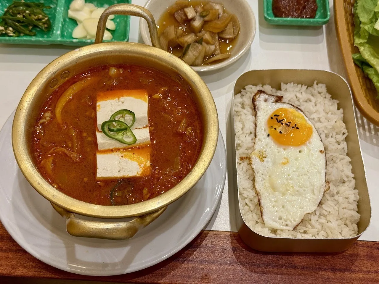 A gold pot of red stew with tofu beside a metal tray of rice with a fried egg