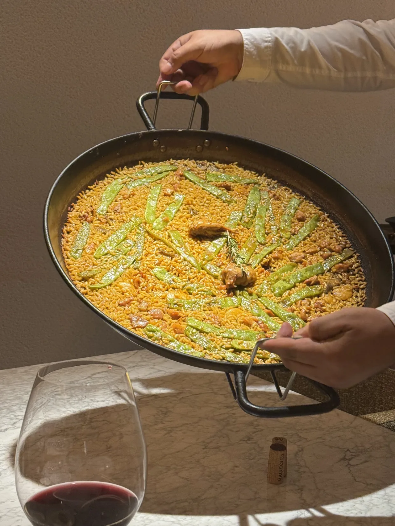 A large pan of yellow rice with green vegetables held over a table, with a glass of red wine