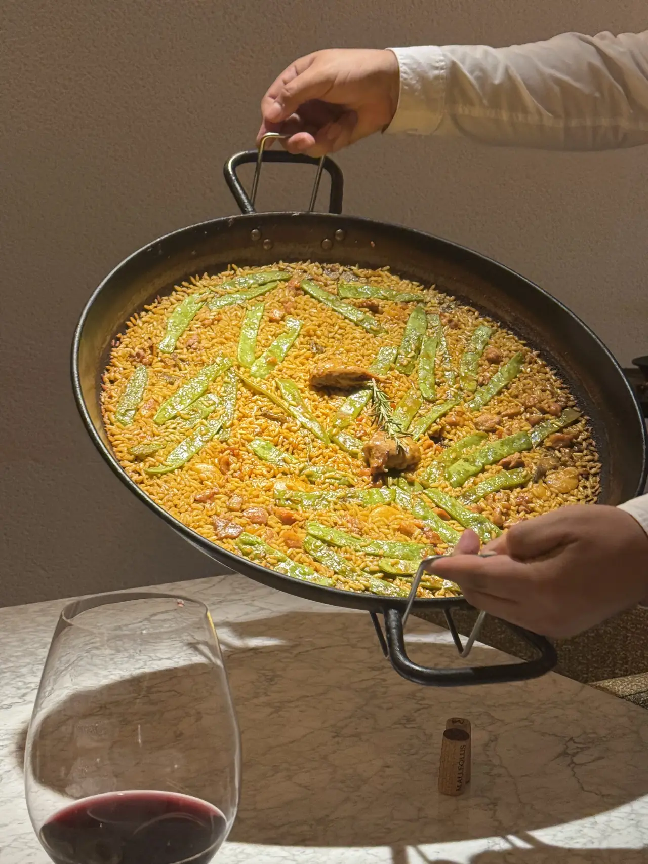 Server presenting a rice dish with runner beans and meat in a paella pan tableside, red wine glass in foreground