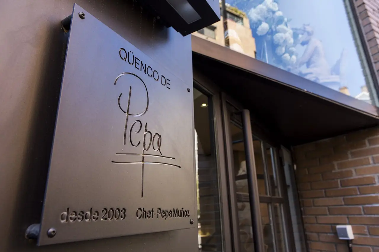 Metal entrance plaque reading Quenco de Pepa, desde 2003, Chef Pepa Munoz, mounted beside the restaurant's glass door and brick facade