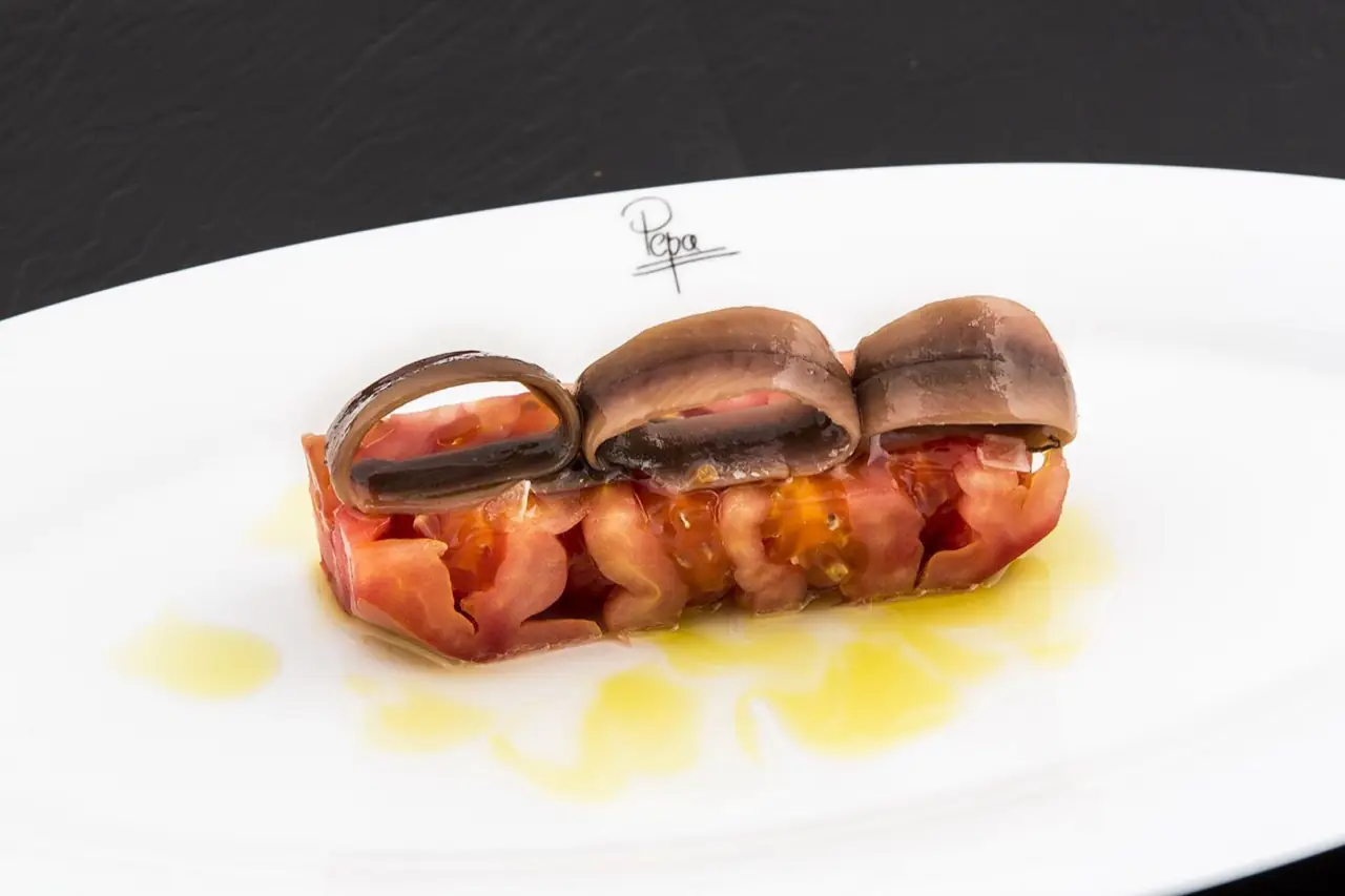 Three rolled anchovy fillets over a bed of chopped ripe tomato dressed with olive oil on a white plate, faint Pepa signature on rim