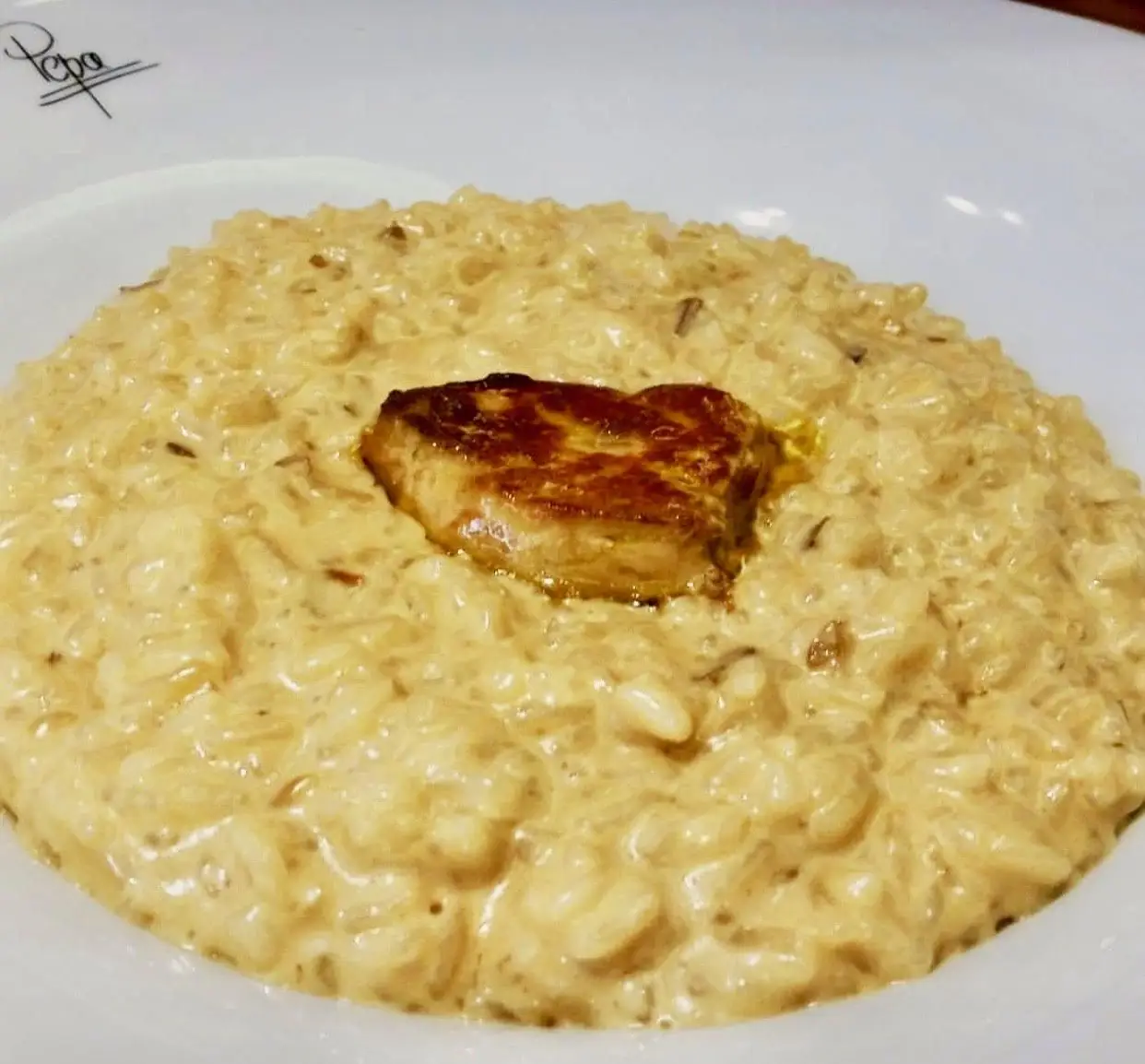 Creamy risotto topped with a seared piece of foie gras on a white plate, faint Pepa signature on the rim