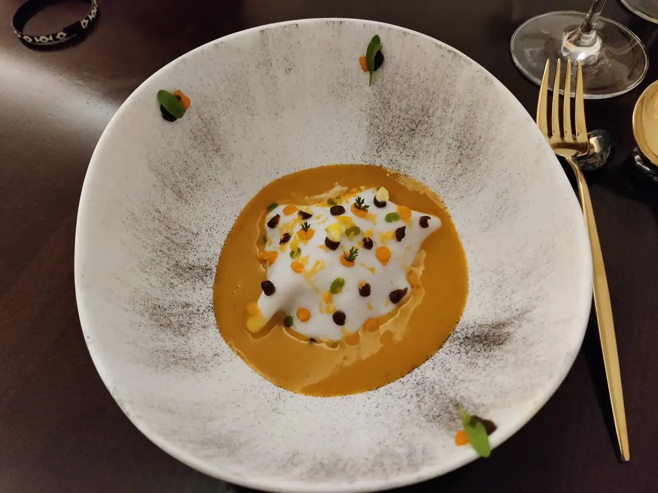 Bowl of orange broth with a foam-topped dumpling-like dish garnished with dots of sauce and herbs