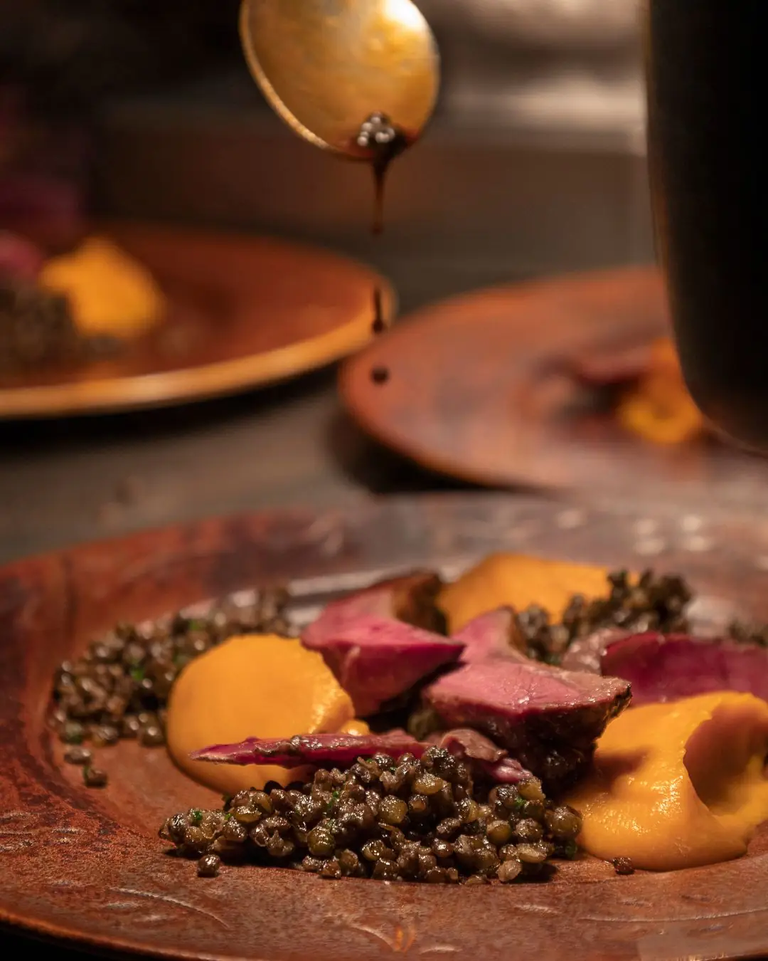 Sauce being spooned over sliced game meat with lentils and squash puree on a plate