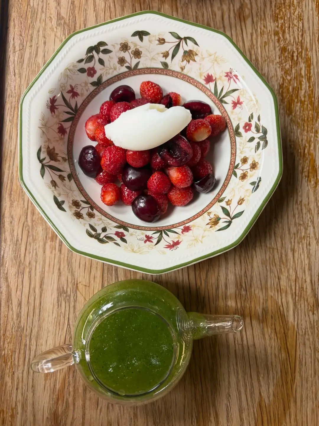 Bowl of strawberries and cherries topped with cream, beside a jug of green sauce