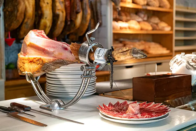 A leg of jamón on a carving stand with a plate of sliced cured meat, next to hanging hams and bread shelves.