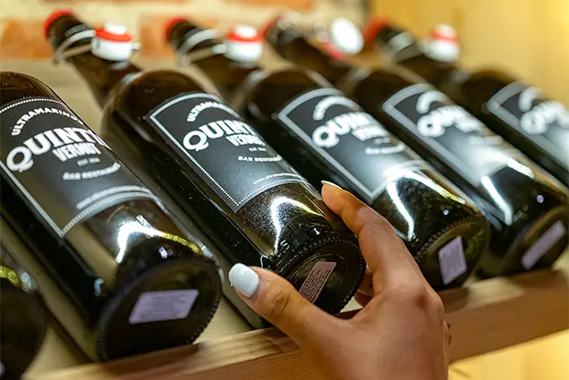 A hand picking a bottle of the shop's own-label vermouth from a row on a shelf.
