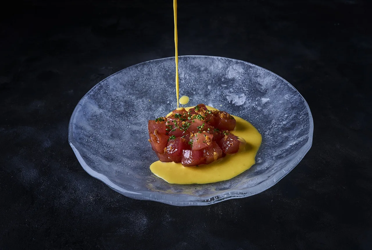 Chopped red ingredient on a yellow sauce in a shallow glass bowl on a dark surface, with a thin stream falling from above