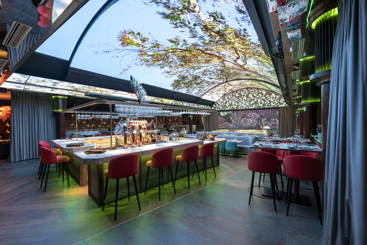 A marble bar counter with red velvet stools and set tables under a domed ceiling screen showing trees