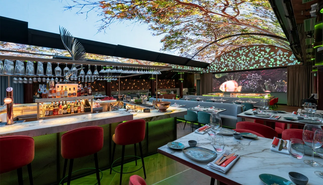 A bar with red stools and set dining tables beside a glass-roofed terrace