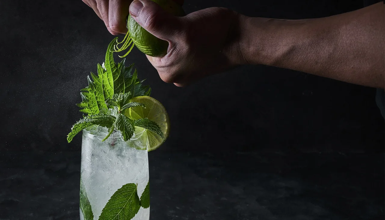 A hand squeezing lime over a mojito with mint on a dark background