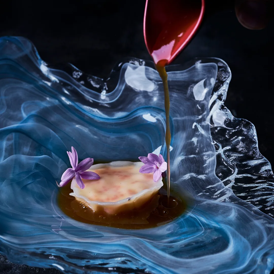 A dish garnished with purple flowers, with sauce being poured over it