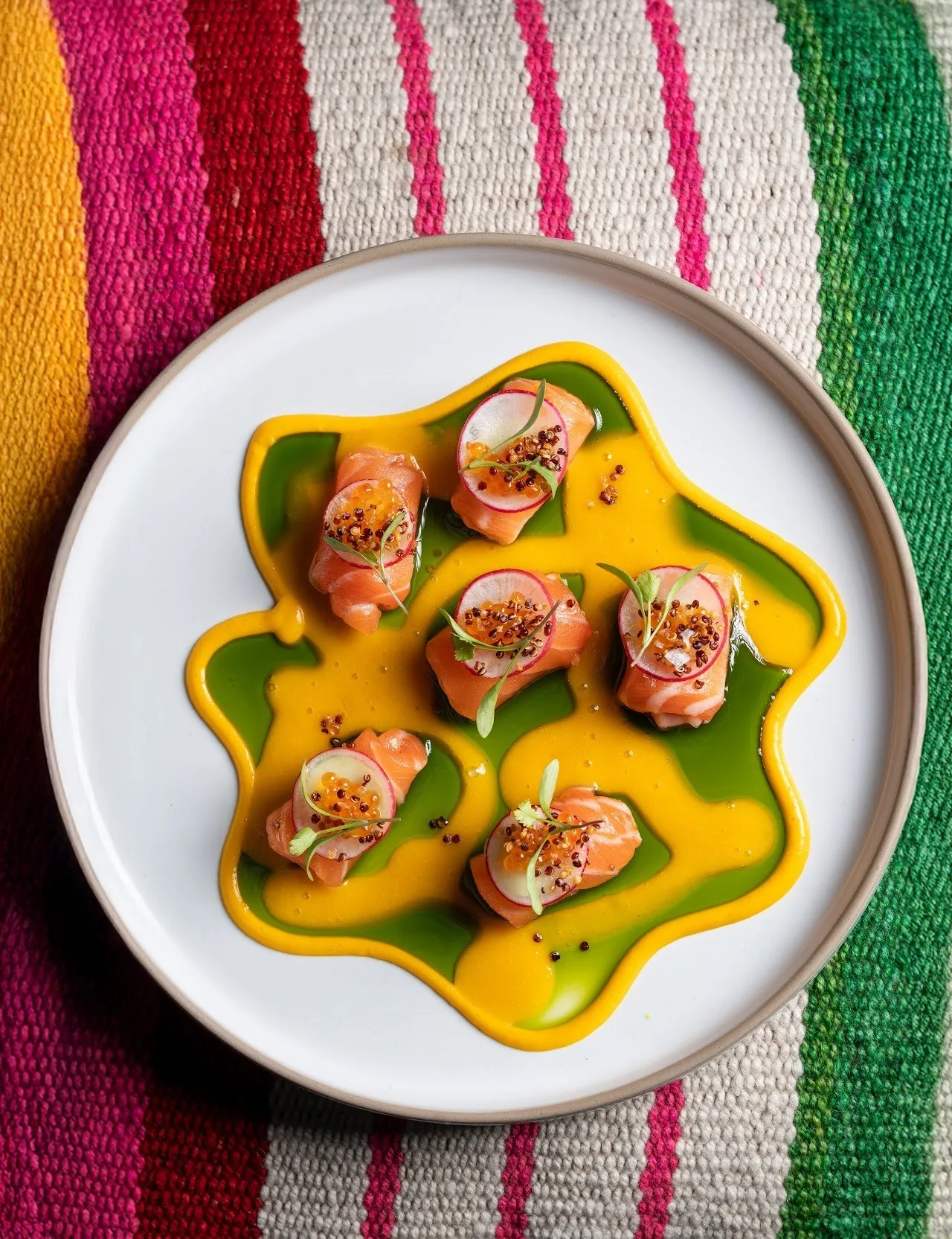 Slices of salmon topped with herbs on a yellow and green sauce on a white plate over a striped textile
