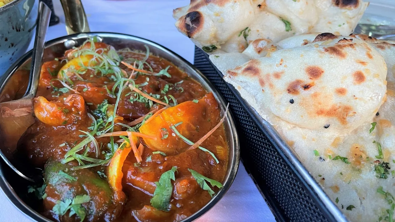 A metal bowl of curry with vegetables and coriander beside a tray of naan bread