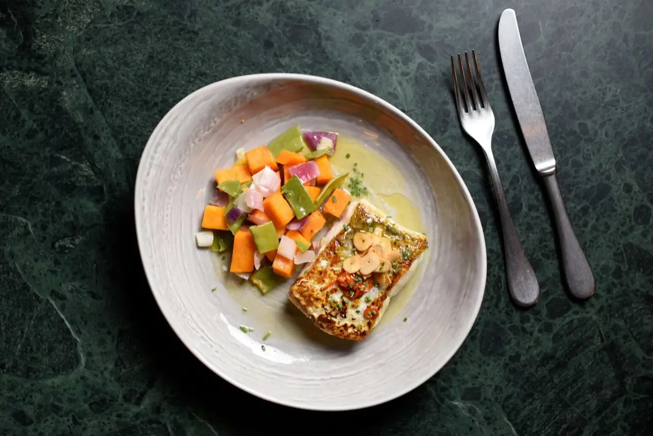 Pan-seared white fish fillet topped with sliced garlic in butter sauce, with diced sauteed vegetables, on a grey plate over green marble