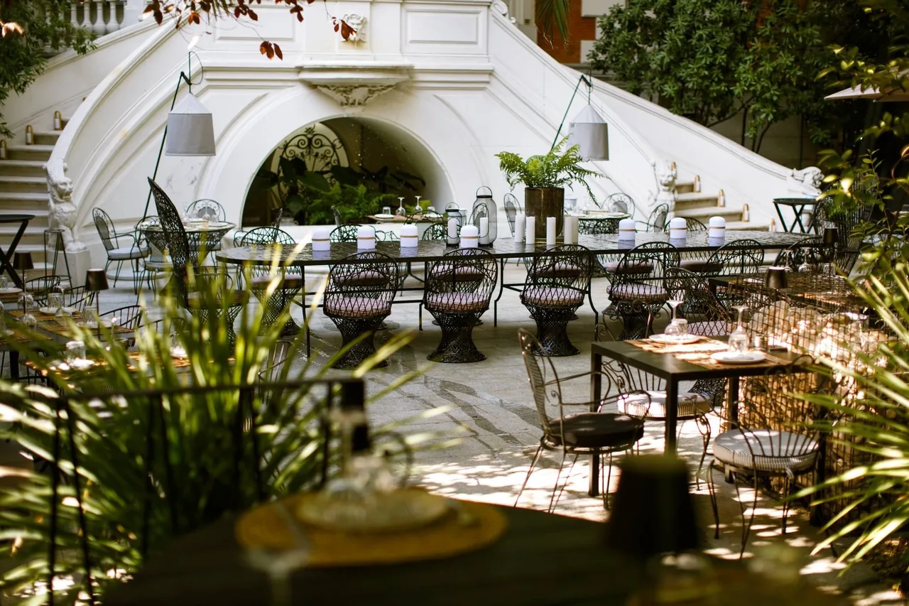 Iron chairs and tables set in a courtyard garden beneath a white stone staircase and plants