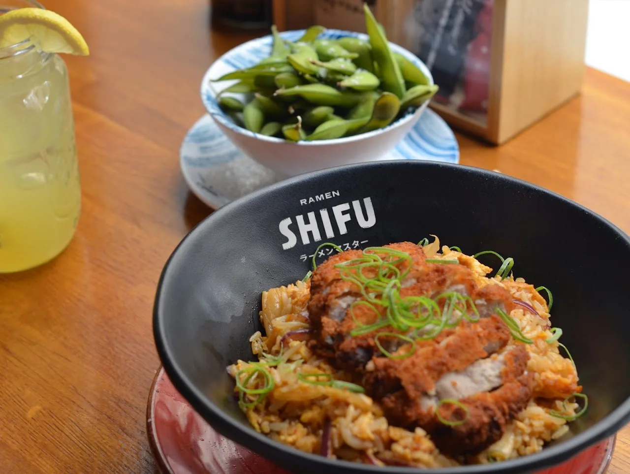 A bowl of katsu rice topped with breaded pork cutlet, next to a bowl of edamame and a glass of lemonade