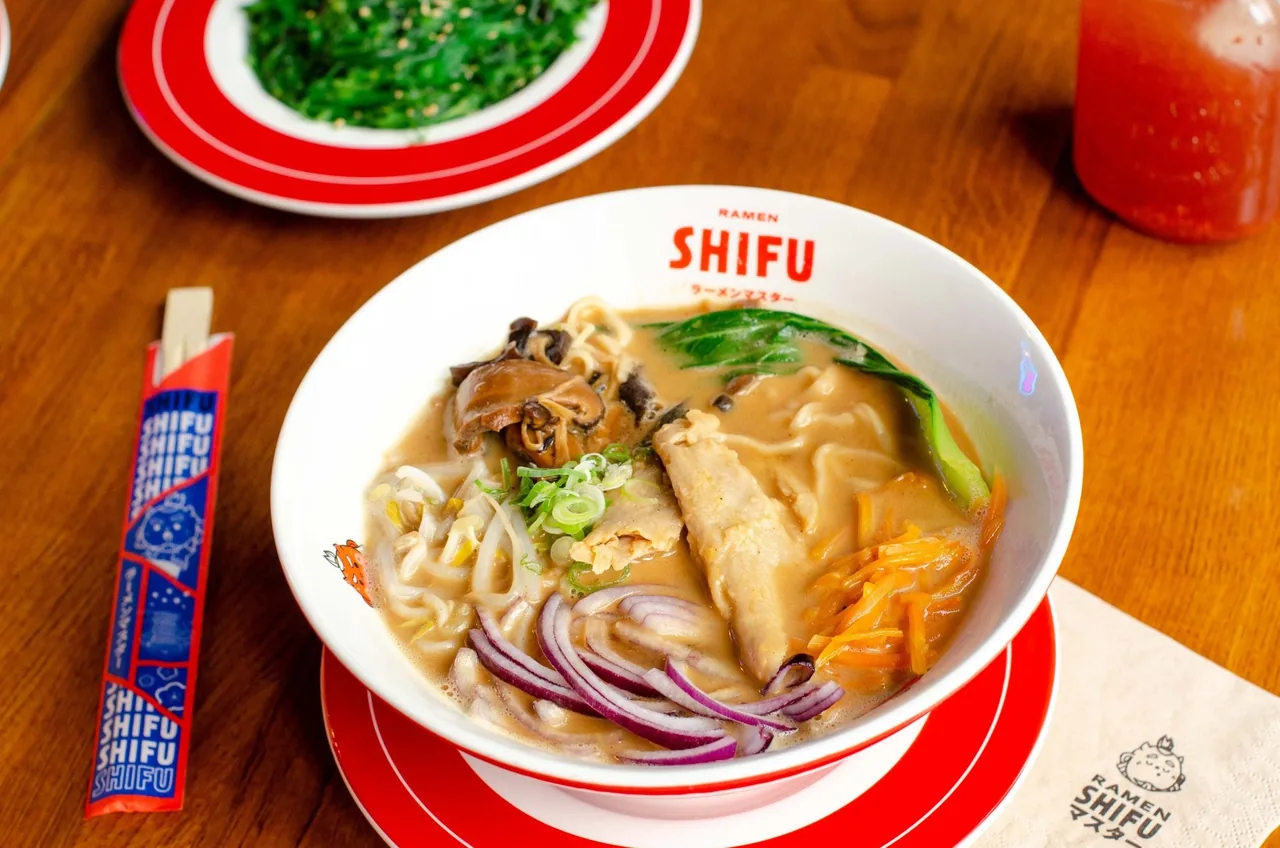 A white ramen bowl with noodles, greens and dumplings in broth on a red-rimmed plate