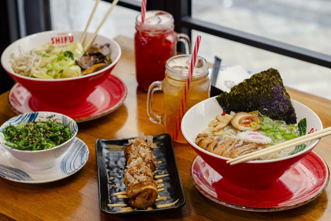 Two bowls of ramen on a wooden table with iced drinks and small side dishes
