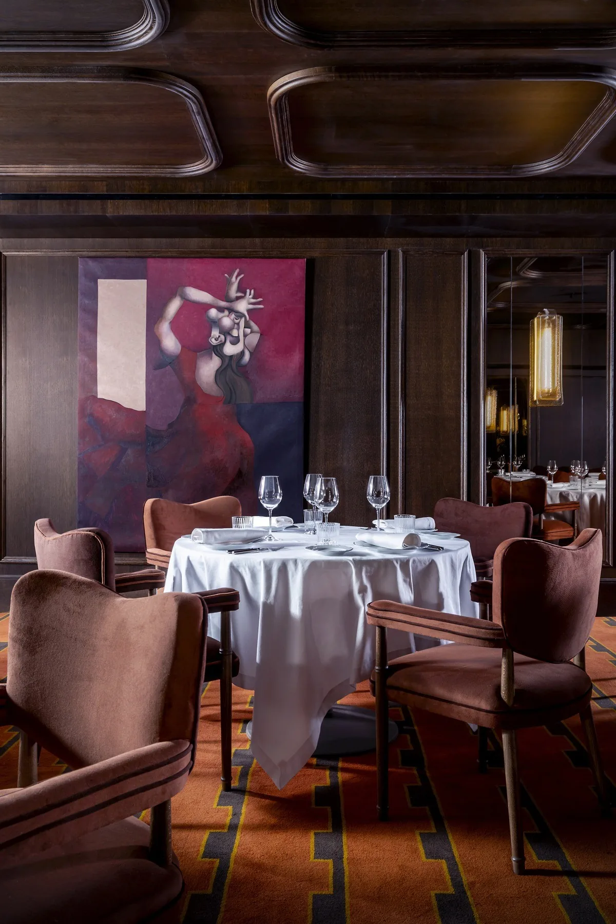 A dining room with a set round table, velvet armchairs, dark wood panelling and a painting on the wall