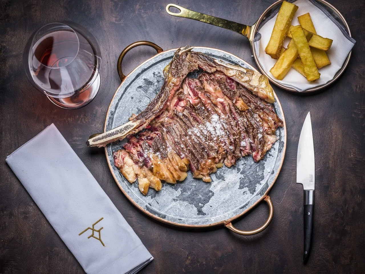 A bone-in steak on a round metal platter with a knife, fries and a glass of red wine