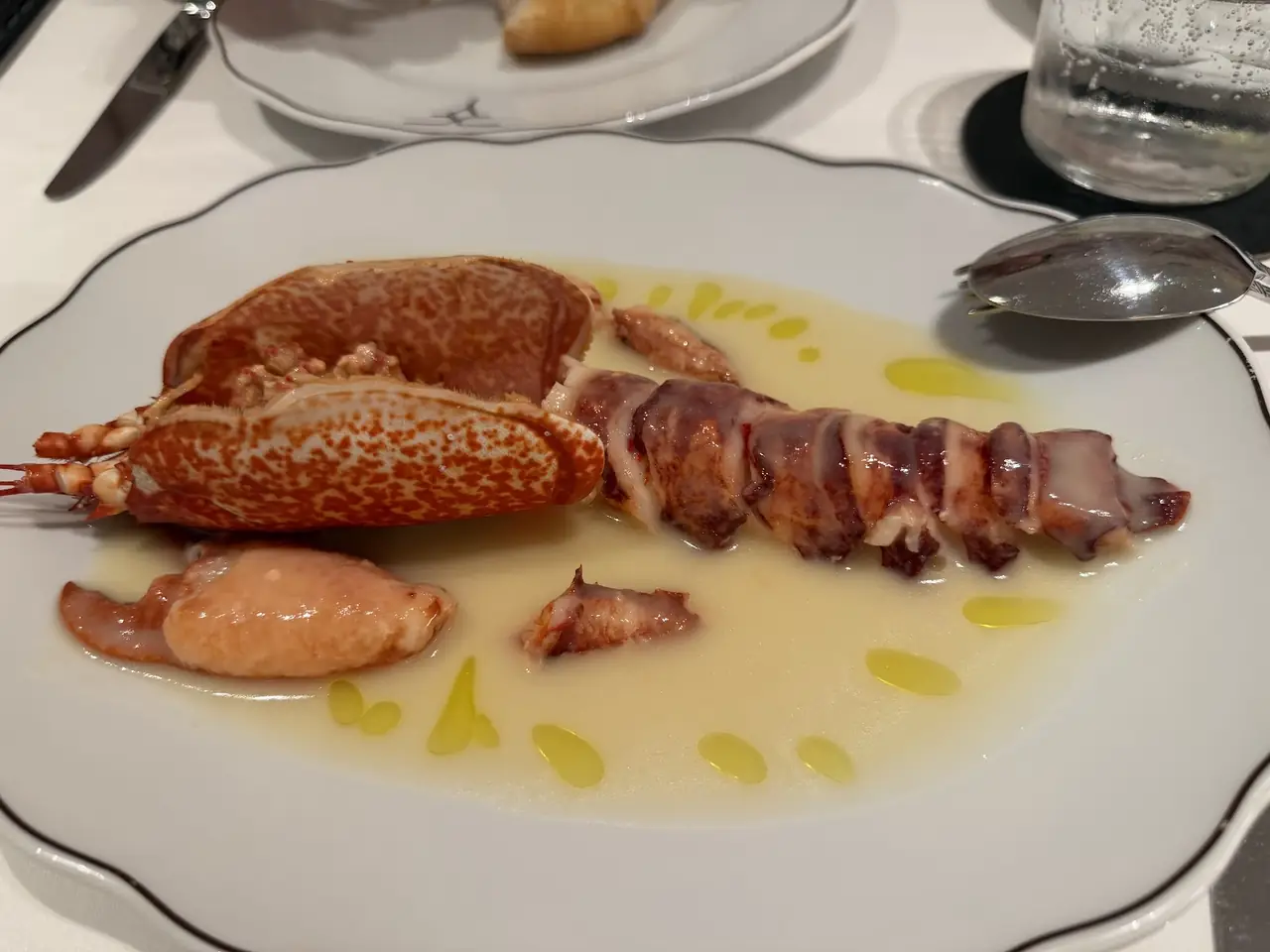 Langoustine tail and head sliced open over a pale butter sauce dotted with olive oil, on a scalloped white plate