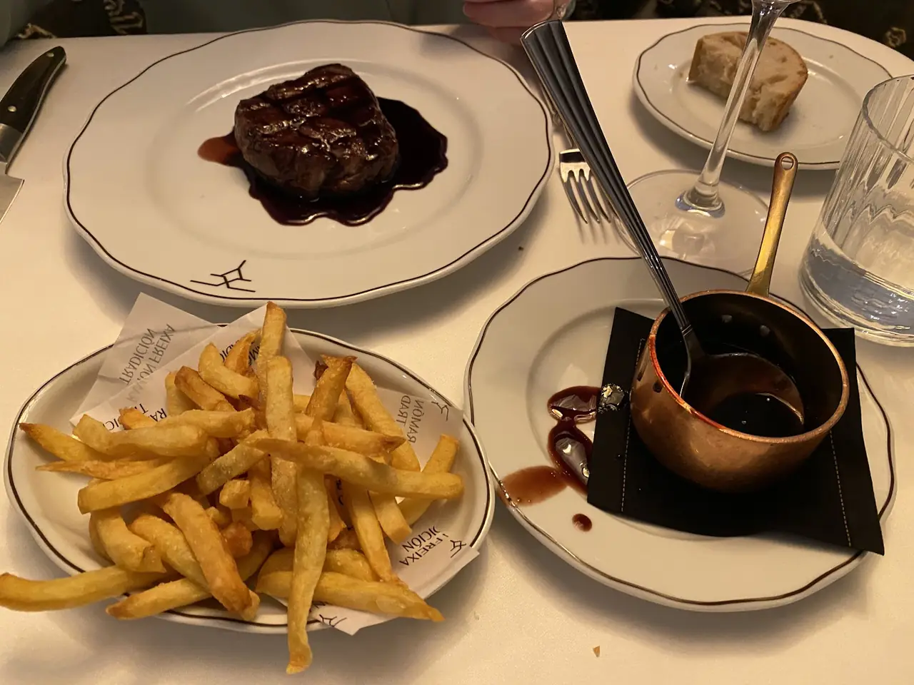 Seared steak filet in a red wine sauce on one plate, a plate of golden fries and a copper pot of sauce alongside on the table