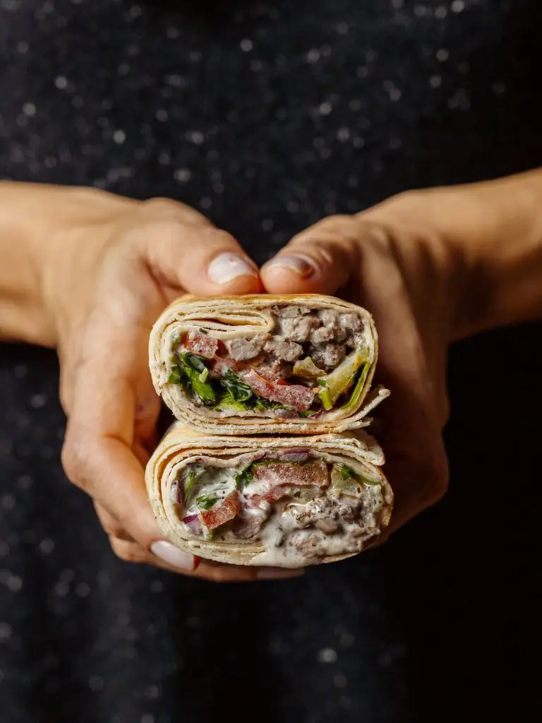 Two hands holding a shawarma wrap sliced to show meat, vegetables and white sauce in cross-section