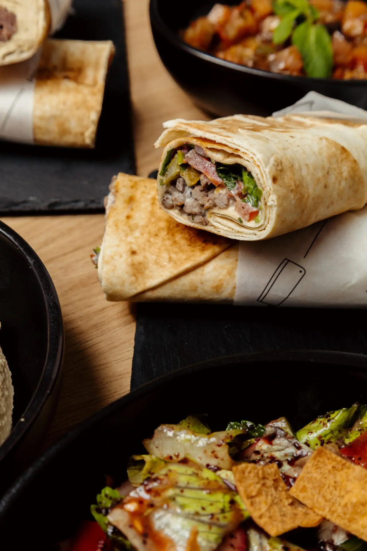 Shawarma wrap cut open to show meat and vegetables, resting on paper wrapping with fattoush and mezze bowls around