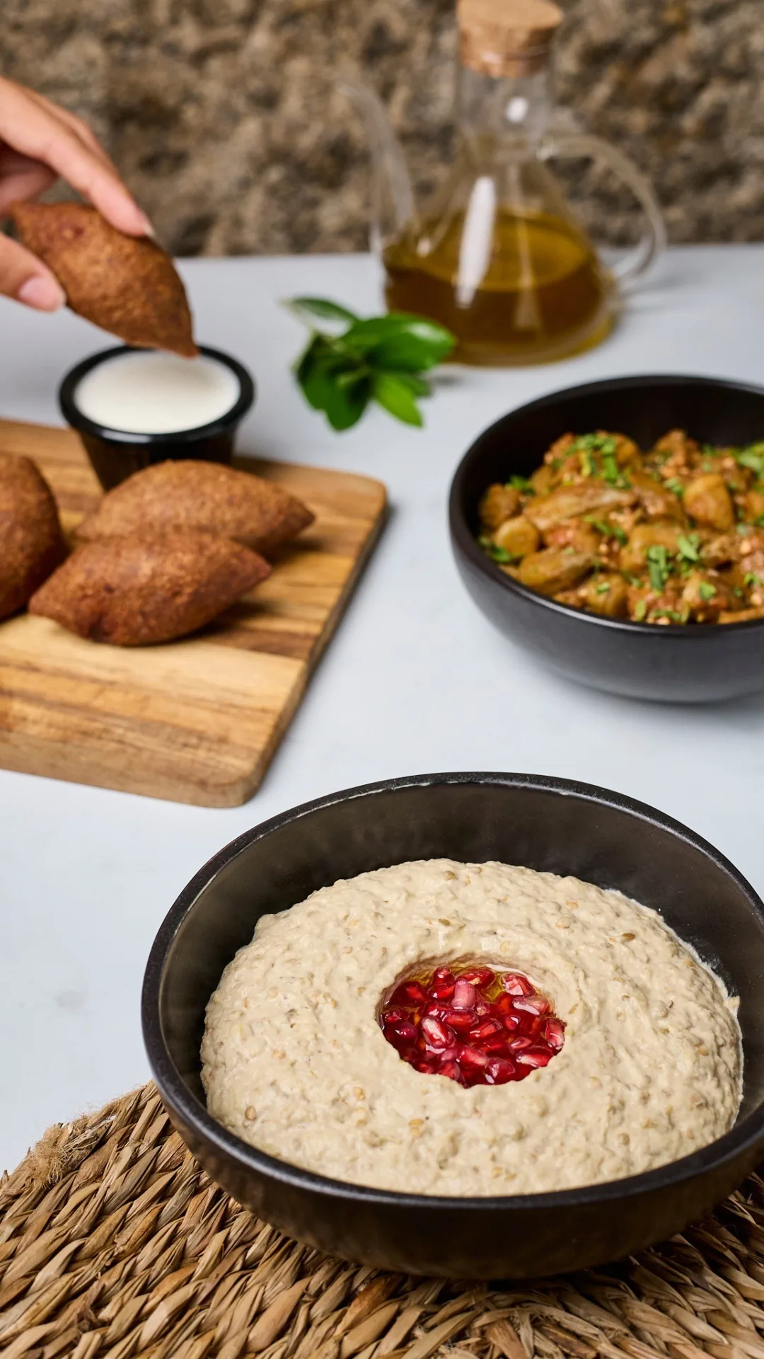 A dark bowl of creamy dip topped with pomegranate seeds beside fried kibbeh on a wooden board