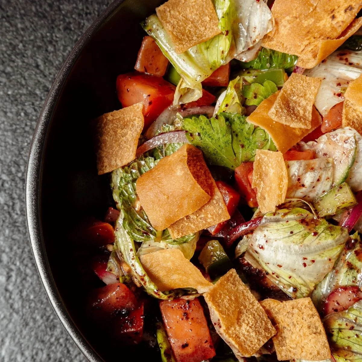 A dark bowl of salad with tomato, lettuce, peppers and pieces of crispy flatbread