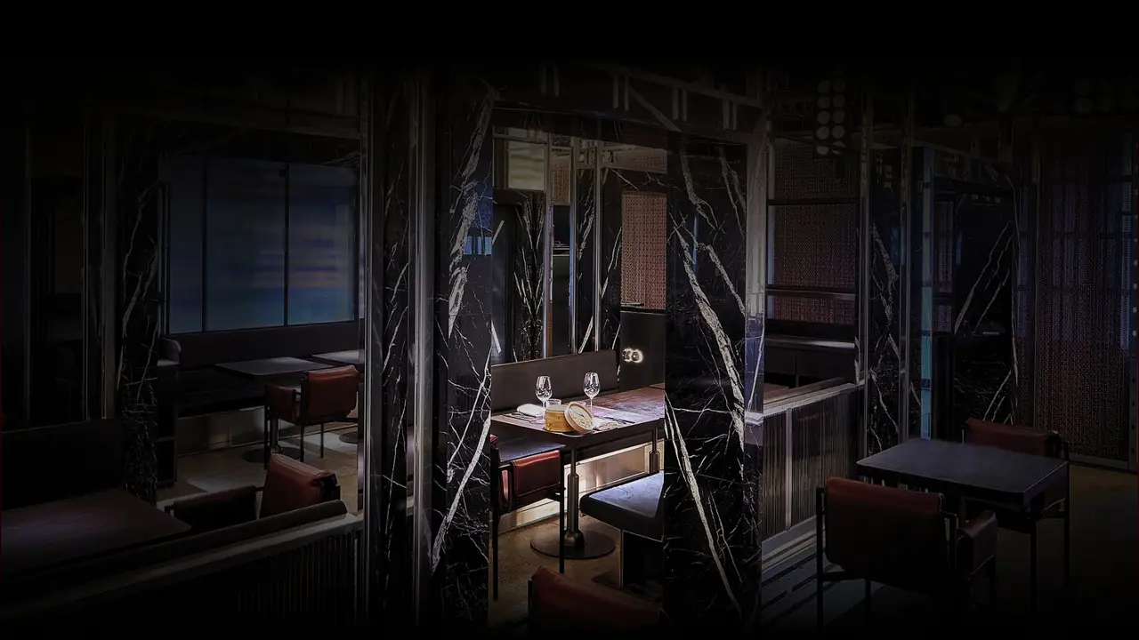 Dark dining room divided by black-and-white marble pillars, a lit booth at the centre with two wine glasses and a stacked bamboo steamer on the table, red leather chairs and a small illuminated XO sign on the wall