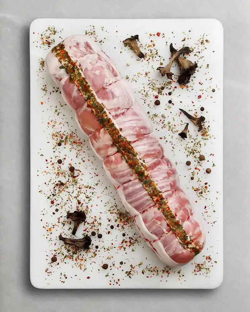 A rolled, seasoned raw pork cut with herb stuffing laid on a white board, garnished with wild mushrooms and peppercorns
