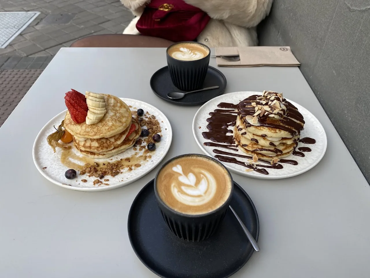 Two plates of stacked pancakes with berries and chocolate drizzle, alongside two coffees