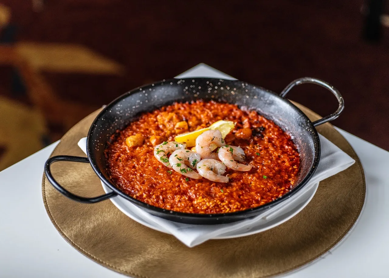 A pan of seafood paella with shrimp and a lemon wedge, in a paella pan on a straw placemat