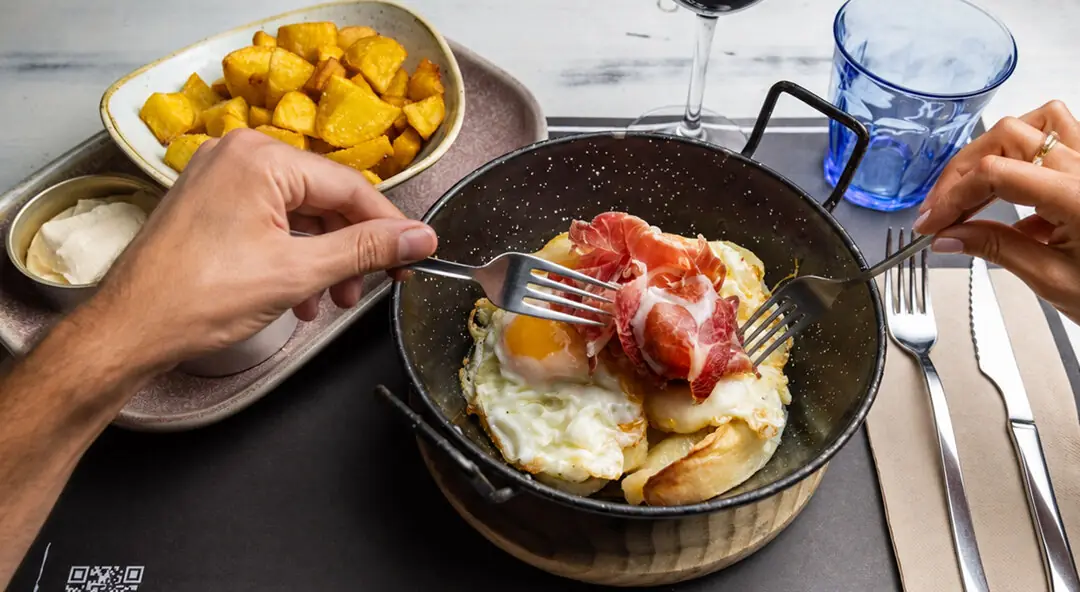 Fried eggs topped with cured ham in a small black pan, served with a side of fried potato cubes, at Restaurant La Pepa